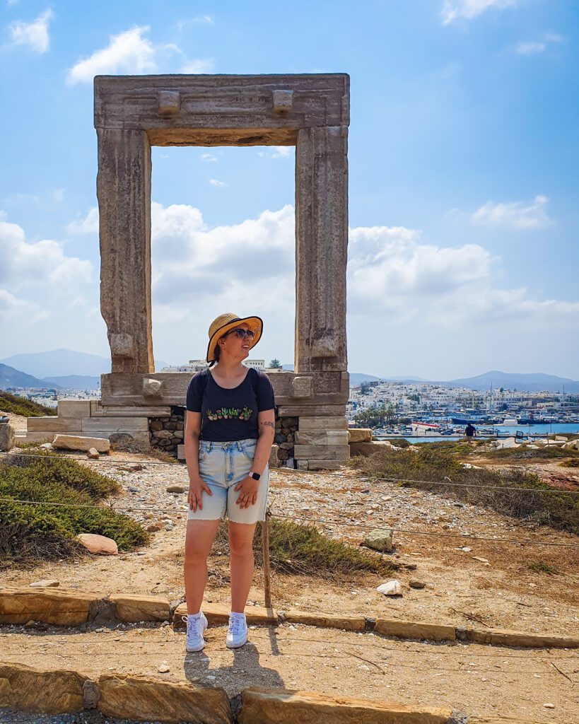 Krissie standing in front of a large stone rectangle which is behind a small barrier. The stone structure is the Portara and is placed on the ground on more large stones and you can see bits of Chora in Naxos, Greece with its white buildings behind it "How to Have the Best Time in Naxos on a Budget" 