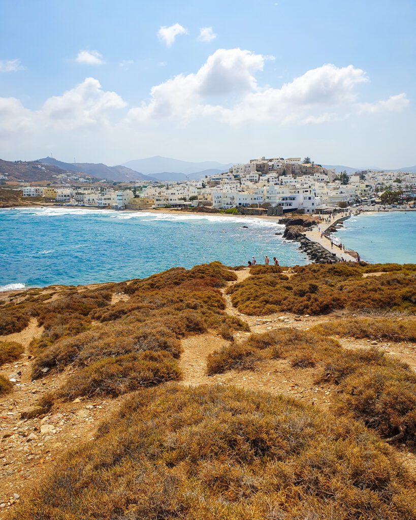 A view back to Chora, Naxos in Greece. In the foreground are lots of brown bushes and walkways through them. In the middle is blue ocean on either side of a walkway across the ocean. The walkway is coming from a town built up over a hill with lots of blocky white buildings "How to Have the Best Time in Naxos on a Budget"