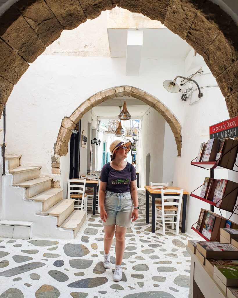 Krissie standing on a cobbled street and under a stone archway in Naxos, Greece. Around her are books on stands from a nearby bookstore, and tables and chairs from a nearby restaurant "How to Have the Best Time in Naxos on a Budget"