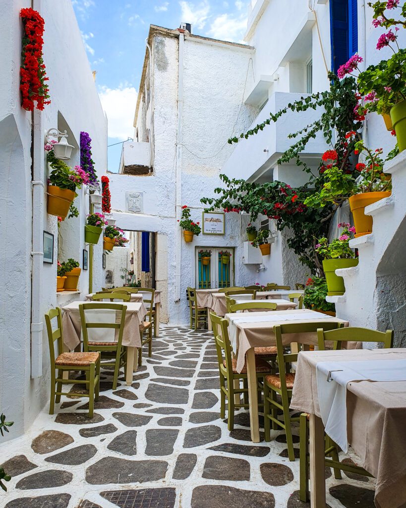 A beautiful view of a restaurant in Naxos, Greece with a white and brown cobbled stone street, white buildings, potted plants and tables and chairs around "How to Have the Best Time in Naxos on a Budget"