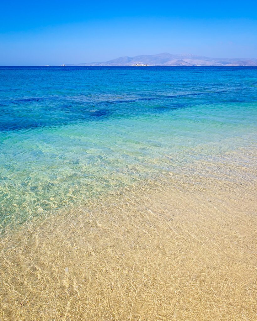 The clearest water we've ever seen, which has a gradient from lighter water to a dark blue further out. On the edge of the water you can see another Greek island "How to Have the Best Time in Naxos on a Budget"