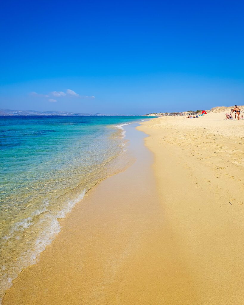 Another beach photo on Naxos, Greece with the water in the left of the photo and a big sand area on the right. There are some people on the sand but it's mostly empty like a lot of the beaches we visited in Greece! "How to Have the Best Time in Naxos on a Budget"