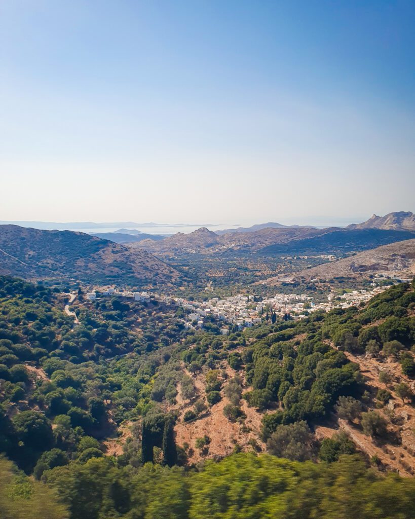 A view over Naxos, Greece with a town of white buildings in the middle. The landscape is covered in green bushes and has lots of hills dotted around "How to Have the Best Time in Naxos on a Budget"