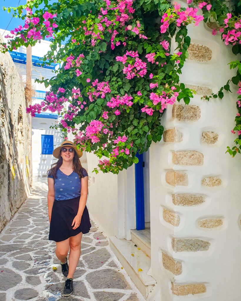 Katherine smiling and walking down the most Greek looking street in Naxos, Greece with its cobbled stone street, white buildings, blue accents on the buildings and a green plant with beautiful pink flowers on it "How to Have the Best Time in Naxos on a Budget"