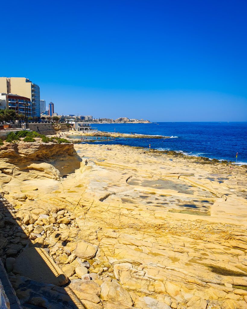 A view along the coast line where you can see some of the cities alone the blue water. Most of the photo is the yellow rock that runs alone the coast beside the water in Malta "Eight Amazing Things to See in Malta"