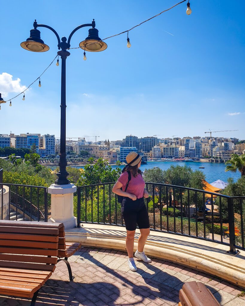 Krissie staring out across a view of a city across the blue water. Beside her is a lamppost and wooden seat and between her and the water are lots of green plants "Where Are the Best Areas to Stay in Malta?"