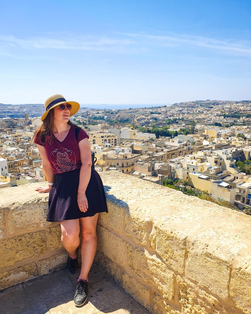 Katherine leaning against a stone barrier with a view of Victoria in Gozo behind her. All the buildings are sand coloured and the land around the buildings is really flat with green bushes "Where Are the Best Areas to Stay in Malta?"