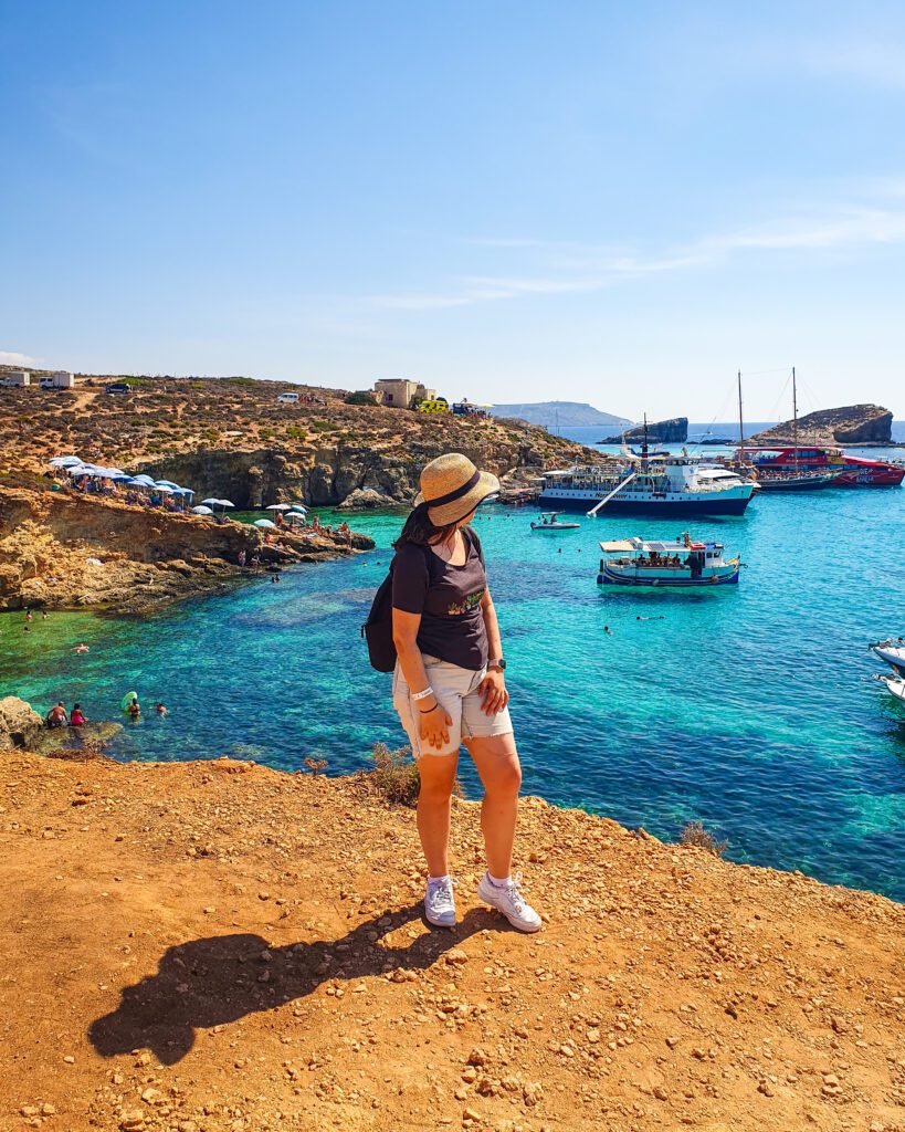 Krissie staring out at the Blue Lagoon on the island of Comino in Malta. The ground is orange and the water is crystal blue. There are also boats and people in the water "Where Are the Best Areas to Stay in Malta?"