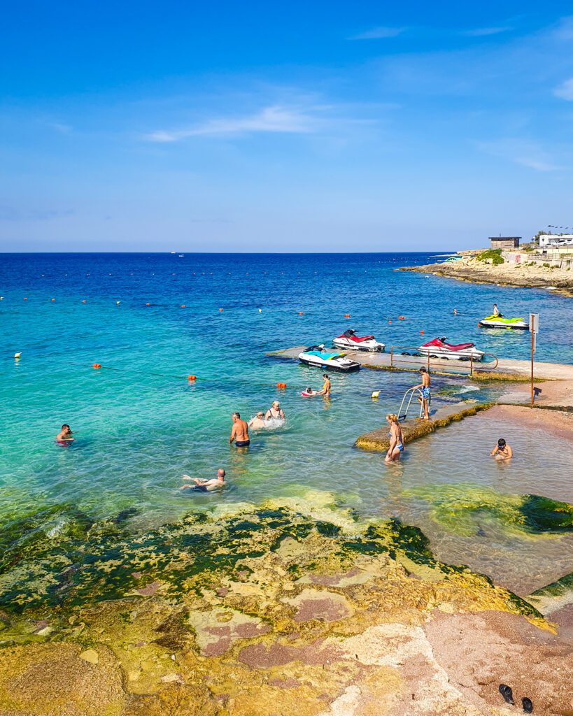 A little beach in St Paul's Bay in Malta with some people swimming in the blue water "Where Are the Best Areas to Stay in Malta?"