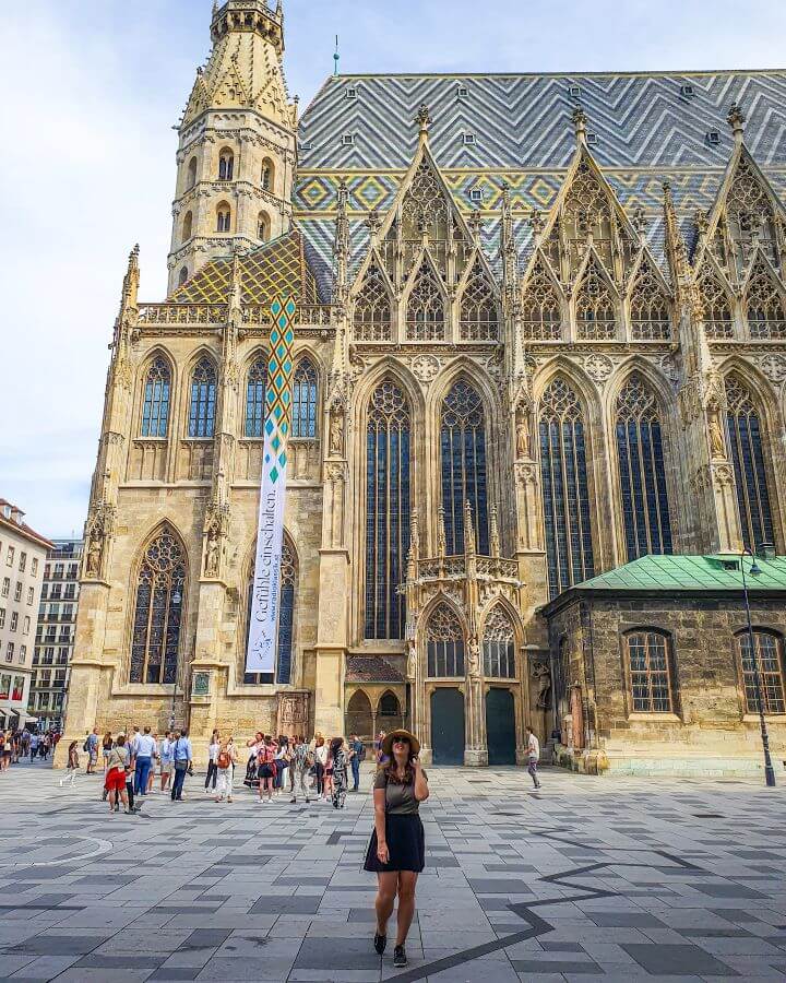 Katherine standing in front of a huge golden cathedral in Vienna, Austria. The cathedral has huge arched windows, pointed parts of the roof while the main roof is tiled in blue, white and yellow tiles "Free and Budget Friendly Ways to Spend Your Time in Vienna"