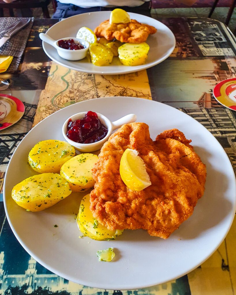 Two white plates with a large golden schnitzel on it with yellow potato halves, a lemon wedge and a little pot of cranberry sauce next to it in Vienna, Austria "Free and Budget Friendly Ways to Spend Your Time in Vienna"