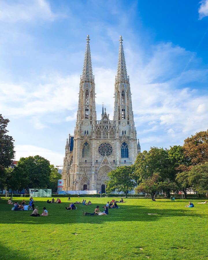 A giant church made of white stone with 2 large spires towering over a large green space with trees and lots of people sitting in the shade "Free and Budget Friendly Ways to Spend Your Time in Vienna"