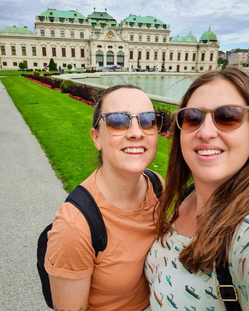 Krissie and Katherine smiling and standing in front of Belvedere Palace in Vienna, Austria. Behind them is a large patch of neatly mown grass and a large white palace with a green roof "Free and Budget Friendly Ways to Spend Your Time in Vienna"