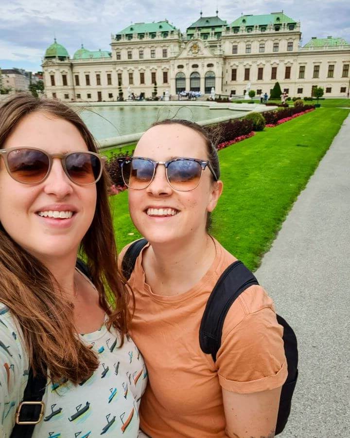 Krissie and Katherine smiling and standing in front of Belvedere Palace in Vienna, Austria. Behind them is a large patch of neatly mown grass and a large white palace with a green roof "Free and Budget Friendly Ways to Spend Your Time in Vienna"