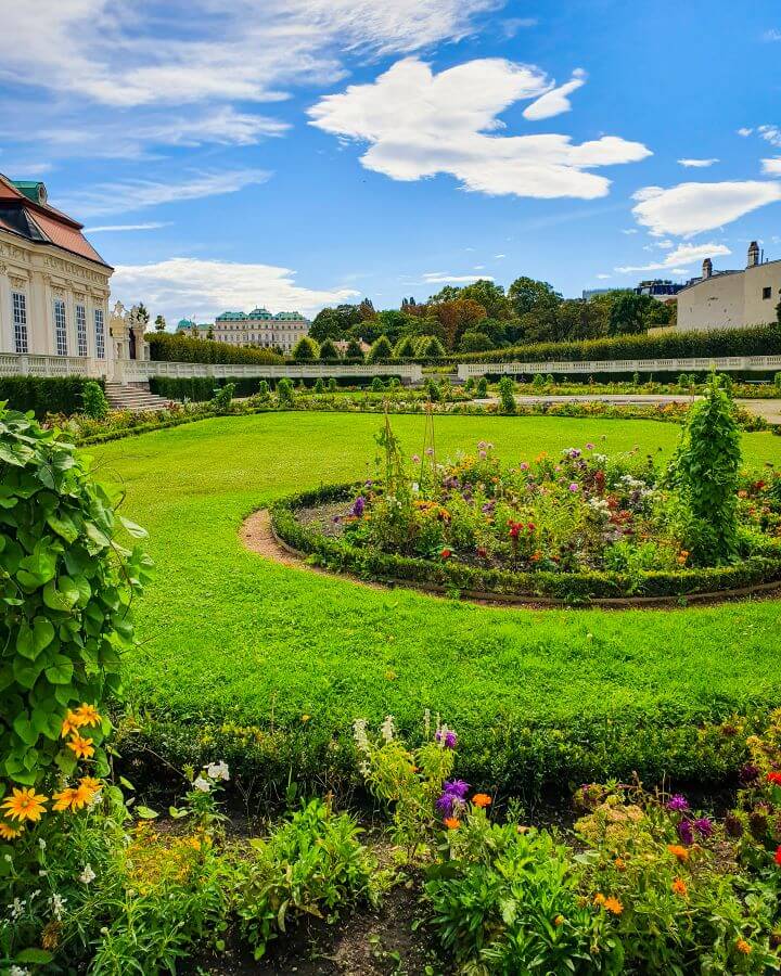 A circular patch of green grass around a flower bed in Vienna, Austria. In the foreground is another flower bed with purple and orange flowers and in the background are green plants and Belvedere Palace "Free and Budget Friendly Ways to Spend Your Time in Vienna"