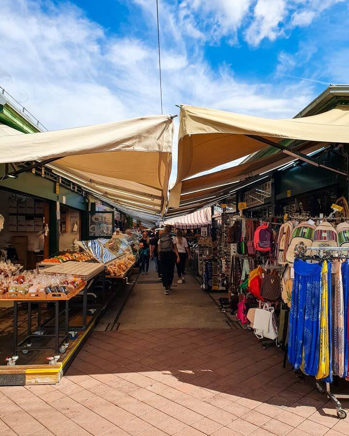 A street with market stalls on both sides in Vienna, Austria and umbrellas covering the whole walkway. Fresh food, clothes, bags and other souvenirs can be seen and there's people walking down the street looking at what's on offer "Free and Budget Friendly Ways to Spend Your Time in Vienna"