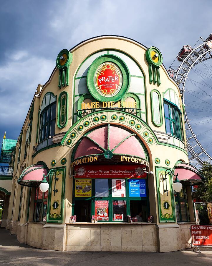 The information desk at Prater in Vienna, Austria which is a building with three red domes over the service windows and a red sign above it that says "Prater". Behind part of a ferris wheel can be seen "Free and Budget Friendly Ways to Spend Your Time in Vienna"