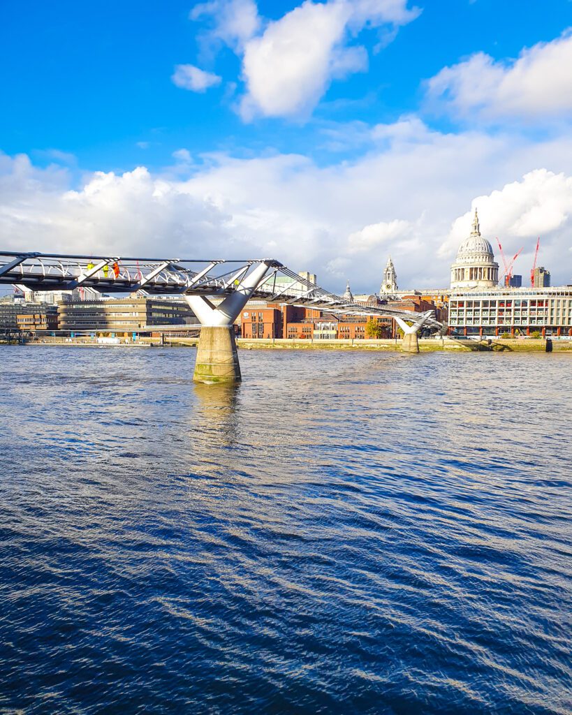 Millennium Bridge going across the Thames river in London, England. The bridge looks a bit like a helix and leads to St Pauls Cathedral on the other side of the river which has a big white dome on top "How to Have the Best Time in London (For Free!)"