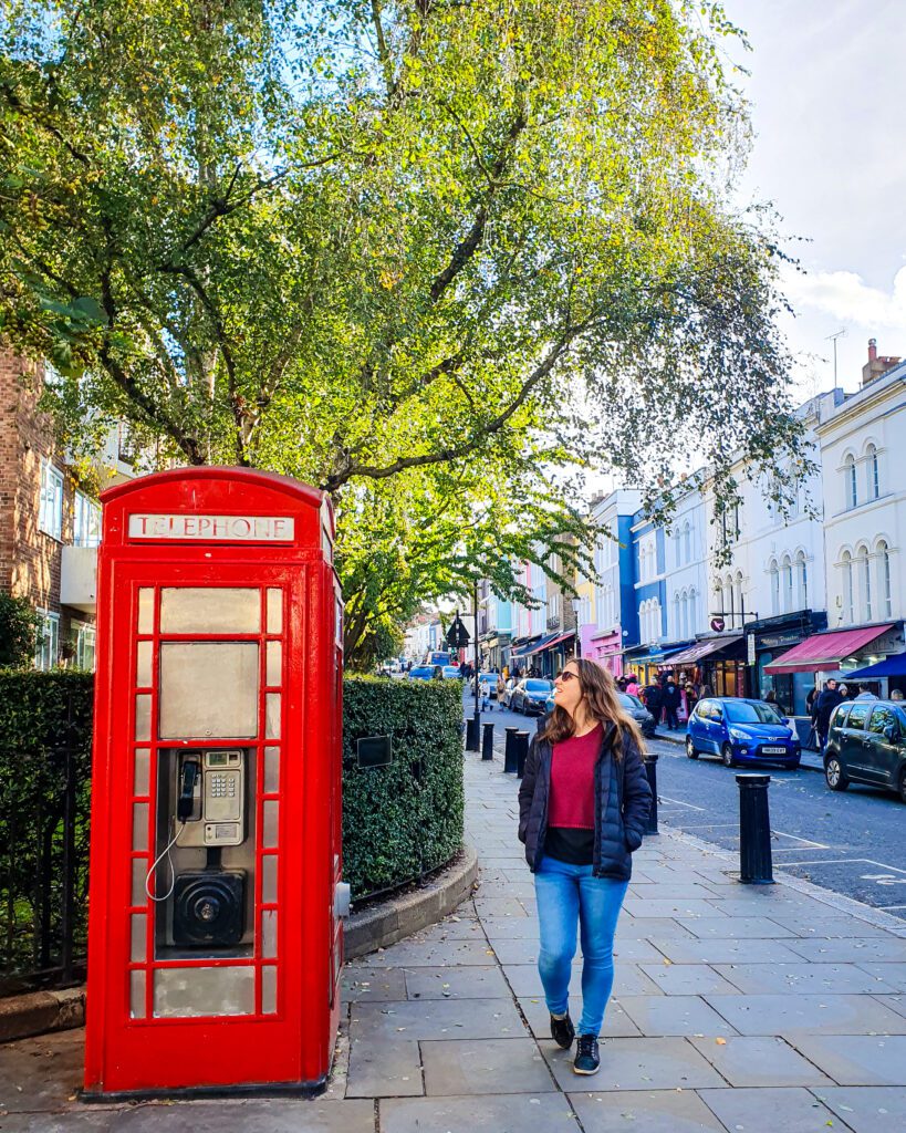 Katherine walking down Portobello Road in London, England and looking to her side at a red telephone box. Behind her is a street with lots of shops, cars and trees "How to See The UK and Ireland Without a Car"