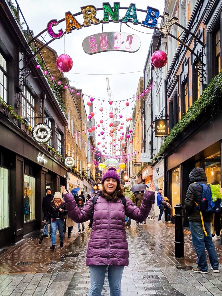 Krissie smiling and putting her hands up to a sign that says "Carnaby Street" in London, England. Behind her is a bustling street with pink and orange decorations strung up between the shops "How to Have the Best Time in London (For Free!)"