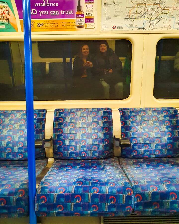 Katherine and Krissie's smiling reflections in the window of the tube in London, England. In front of the window are 3 empty chairs with blue fabric on them "Three Great Hacks To Save Money In London"