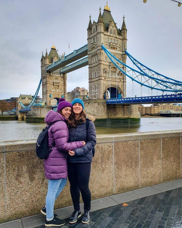 Krissie and Katherine hugging and smiling with puffer jackets and beanies on. Behind them is tower bridge in London, England which has 2 giant stone towers with blue metal strung between them "Navigating the Schengen Area: What You Need To Know (Free Tracker Included)"