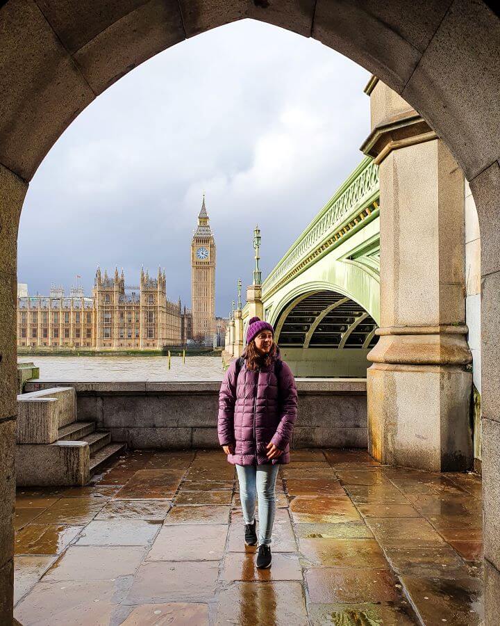 Save Money In London. Krissie smiling and standing under an archway with The Houses of Parliament, Big Ben and the Westminster Bridge behind her in London, England 