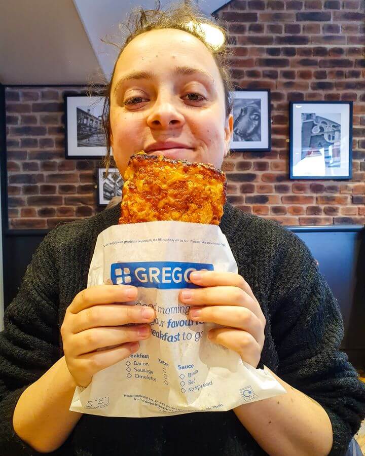 Krissie smiling and holding a paper bag that says "Greggs" with the top of a square pizza coming out of it. Greggs are everywhere in England! "Three Great Hacks To Save Money In London"