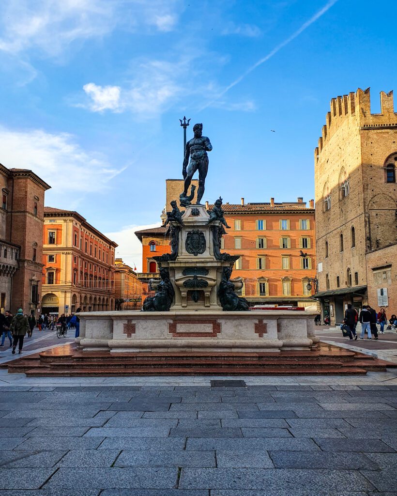 A statue in a town square in Bologna, Italy. The statue is made of stone with bronze figures on it including a big one on the top in the middle of Neptune holding a trident. Around the statue are some orange and yellow buildings "How to Spend Your Time in Verona on a Budget"