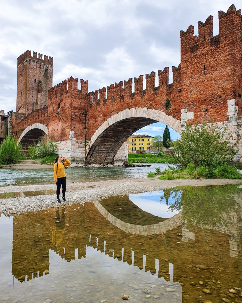 Krissie standing on a rock bed in the Adige river in Verona, Italy. Behind her is the Castelvecchio bridge which is a big red brick bridge with castle like architecture and a turret on the left side. There's also two large arches for the river to go under and some plants around the bridge "How to Spend Your Time in Verona on a Budget"