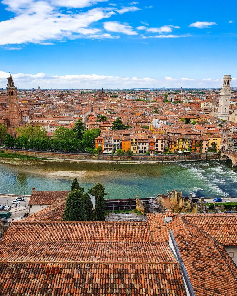 Another view of Verona, Italy from Castel San Pietro. In the foreground are some tiled roofs, the Adige river and beyond that is the coloured buildings and red roofs of the city. On either side of the photo is a big bell tower, one made of brick with a pointed roof and one made of white stone "How to Spend Your Time in Verona on a Budget"