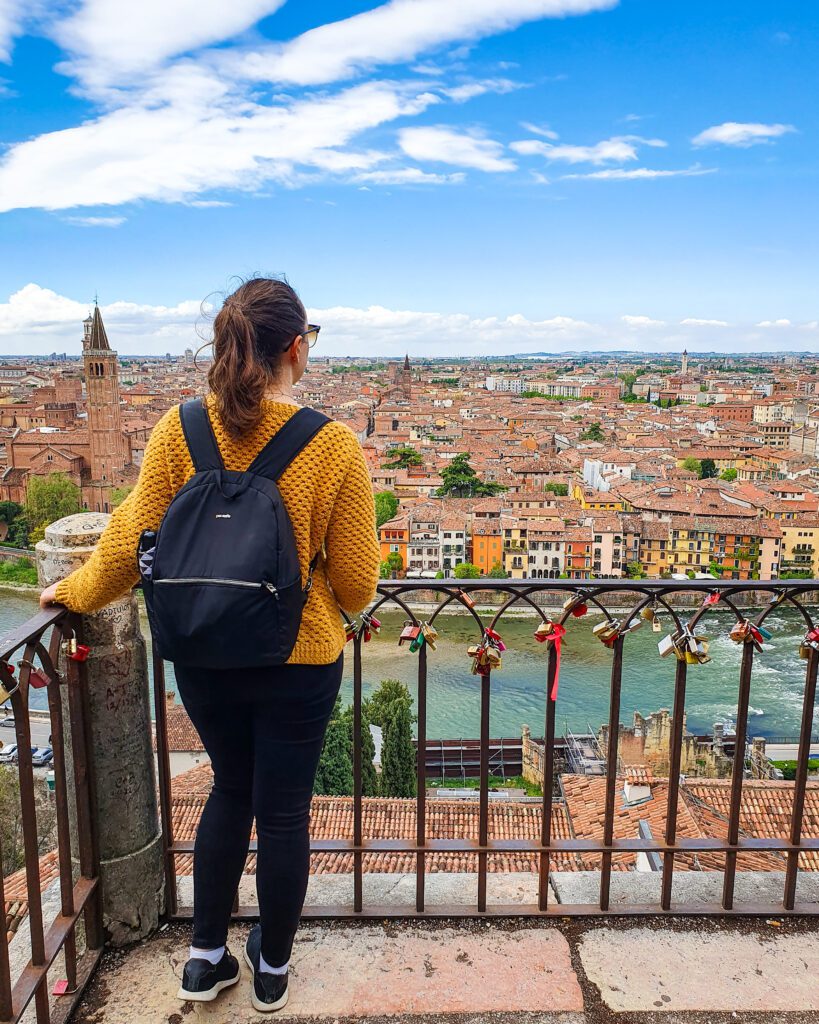 Krissie with her back to the camera and standing in front of a metal railing. Beyond the railing is a sweeping view of Verona, Italy with lots of coloured buildings with red roofs and a church with a big bell tower with a pointed roof. Between the viewpoint and the city is the Adige river "How to Spend Your Time in Verona on a Budget"