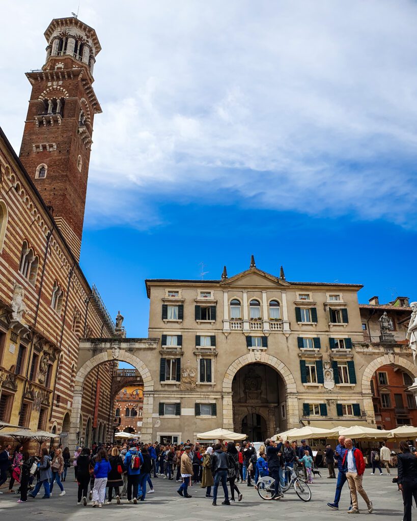 A town square in Verona, Italy busy with people walking around and a big bell tower on the left made of dark stone. In the middle is an old looking building with 3 large stone arches  "How to Spend Your Time in Verona on a Budget"