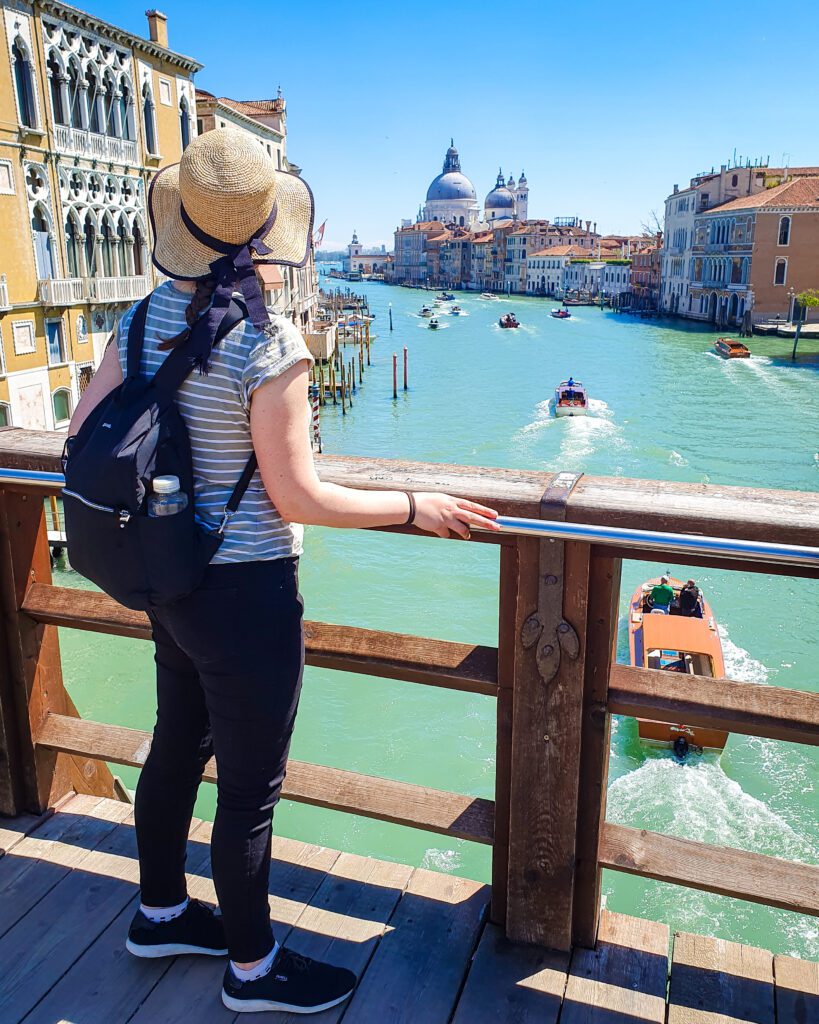 Krissie facing away from the camera standing at a wooden railing. Beyond the railing is a large canal in Venice, Italy with lots of boats on it. Around the water are orange and yellow buildings and in the distance is a big building with two black domes "How to Spend Your Time in Verona on a Budget"