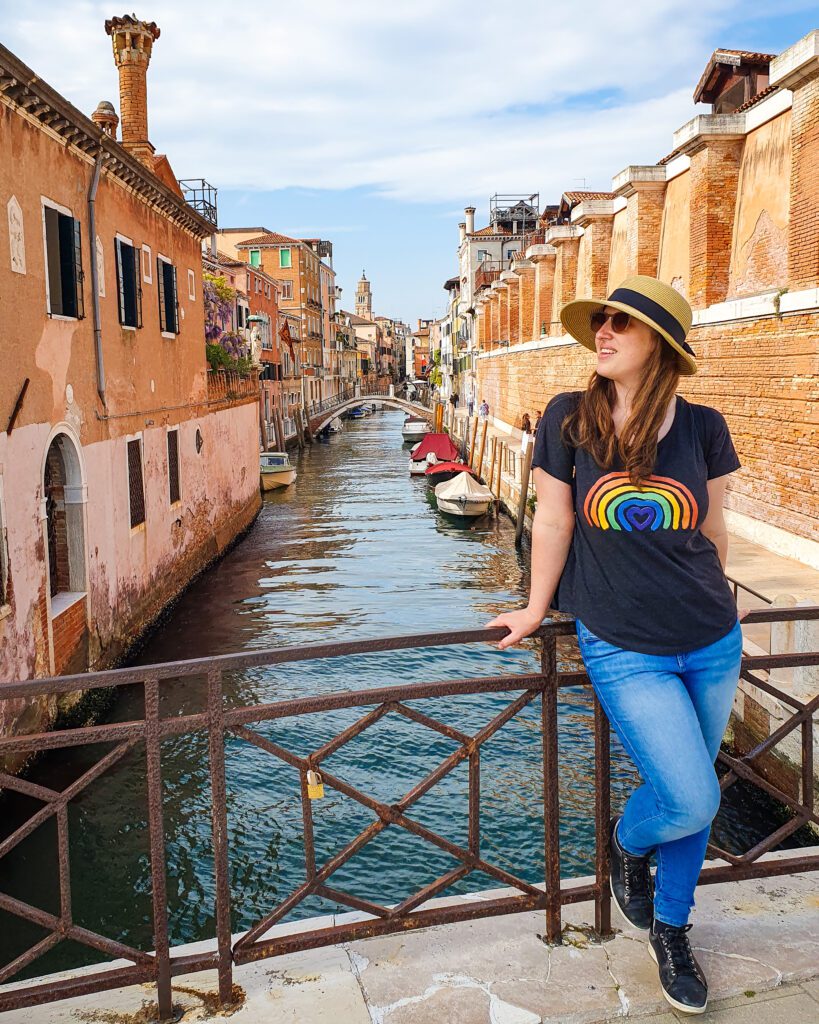 Katherine smiling and standing in front of a metal railing with a canal behind her in Venice, Italy. Around the water are orange buildings with a little stone tower. On the water are some boats and in the middle of the photo is a little bridge going across the canal "How to Spend Your Time in Verona on a Budget"