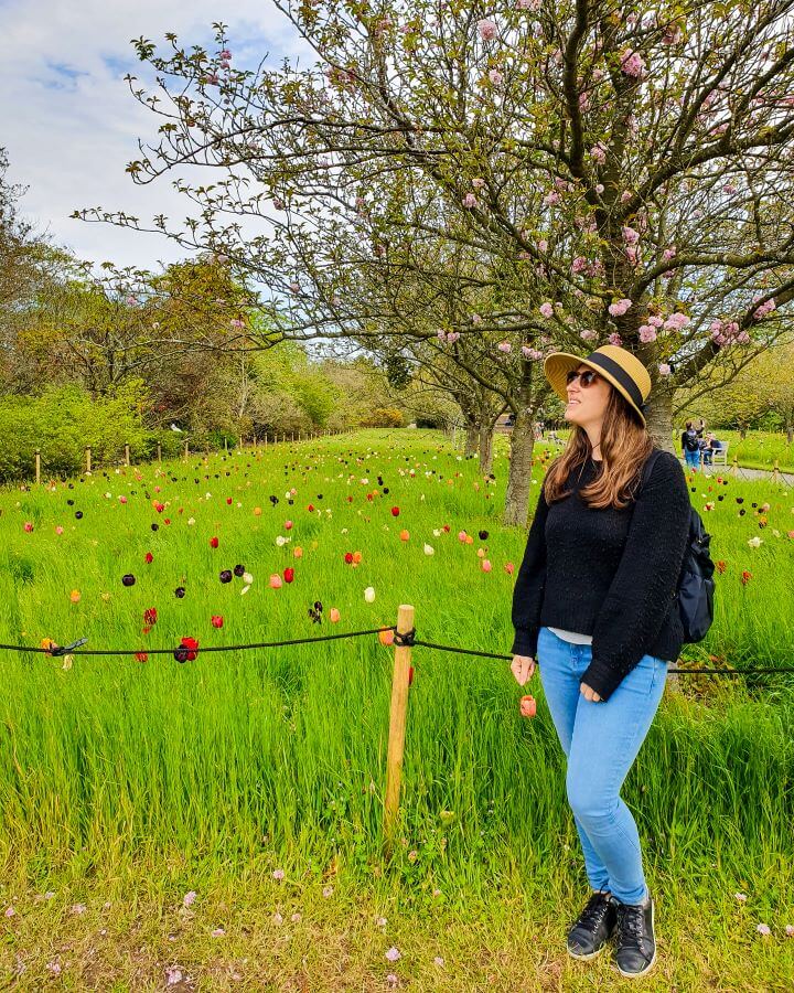 Katherine smiling and standing in front of a field of grass and flowers as well as some trees with blossoms in Kew Gardens in London, England "Three Great Hacks To Save Money In London"