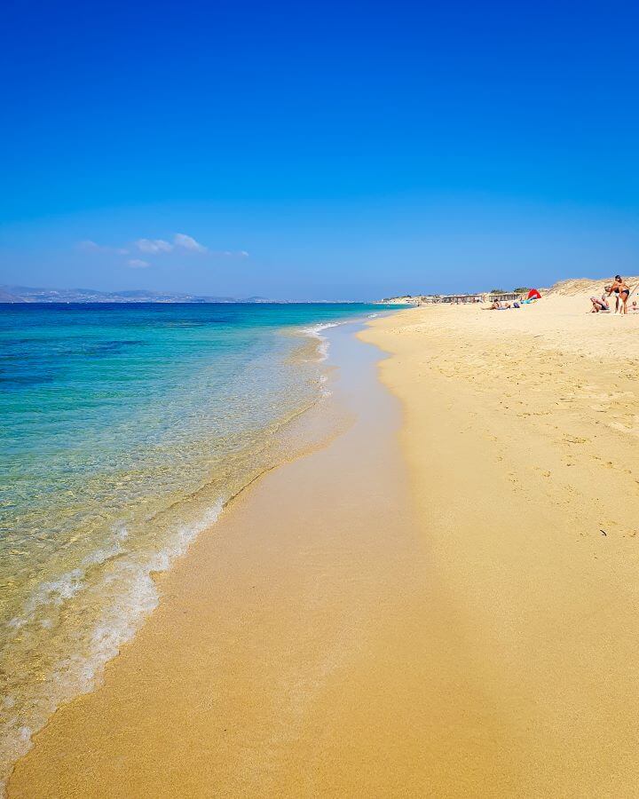 Another beach photo on Naxos, Greece with the water in the left of the photo and a big sand area on the right. There are some people on the sand but it's mostly empty like a lot of the beaches we visited in Greece! "How to Have the Best Time in Naxos on a Budget"