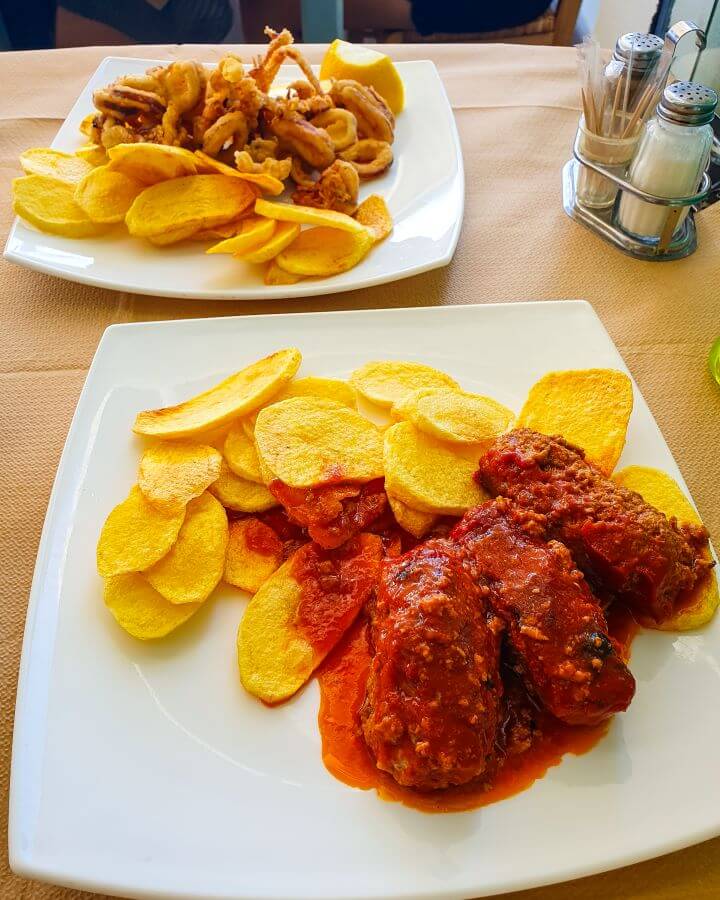 Two plates of food in Naxos, Greece. One has sliced and fried potatoes with fried squid and the other has the same sliced and fried potatoes with 3 large meatballs on it "How to Have the Best Time in Naxos on a Budget"