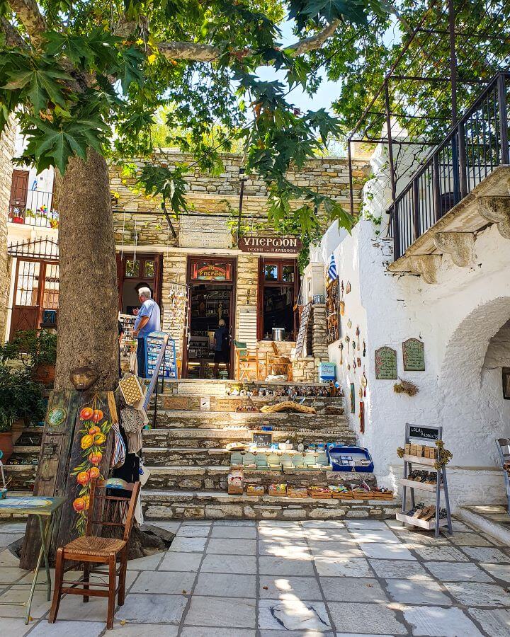 A little square in Apeiranthos, Greece where the floor is made of marble and there are some stone steps up to a little shop. There's a tree on the left and a white building on the right "How to Have the Best Time in Naxos on a Budget"