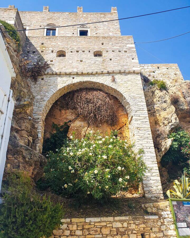 A large stone building in Apeiranthos, Greece built on a rock with a large stone archway that has a big green plant with white flowers at the bottom of it "How to Have the Best Time in Naxos on a Budget"