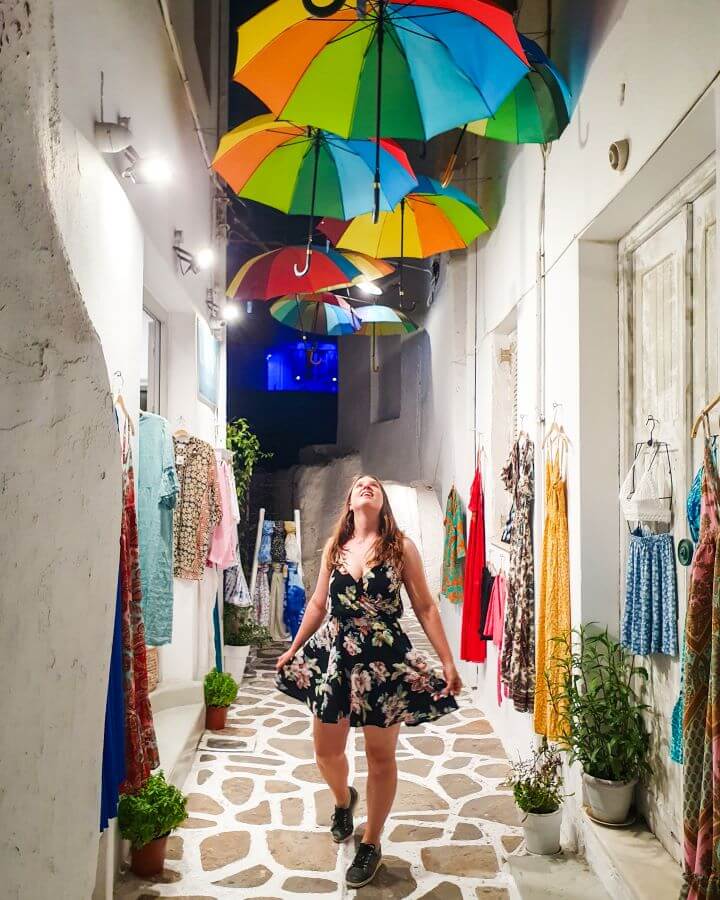 Katherine standing on a cobbled stone street in Naxos, Greece and looking up at some rainbow umbrellas. Around her are some colourful clothes from a nearby shop hanging on the walls of the street "How to Have the Best Time in Naxos on a Budget"