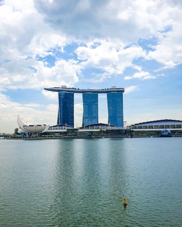 A picture of the Marina Bay Sands hotel in Singapore that has 3 skyscrapers with a long platform across the top of all three. In front of the hotel are more buildings including one that looks like a white flower opening and a big stretch of water in the bay "Singapore: A Complete Guide to the Public Transport System"