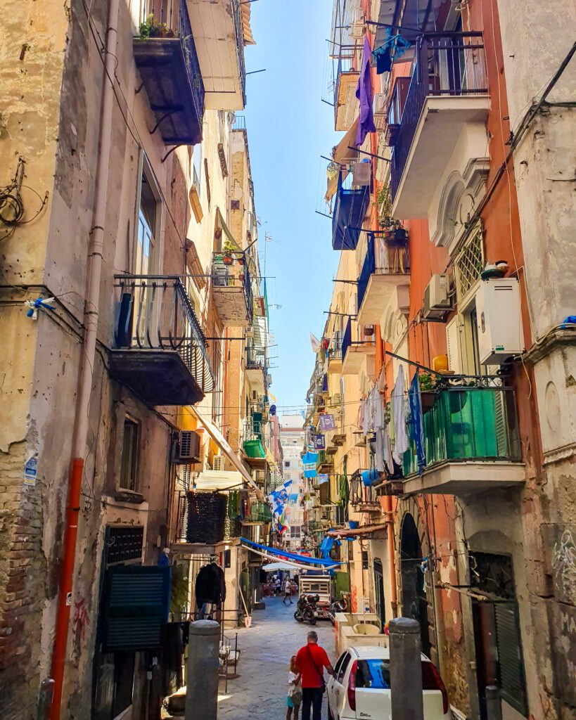 A crowded and narrow street in Naples, Italy with heaps of little balconies with washing hanging off them "See Naples On a Budget With These Six Great Ideas!"