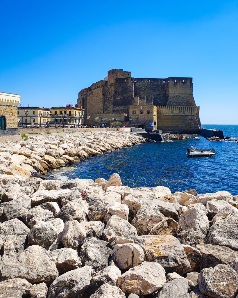 A view along the blue waterline with lots of large rocks along the water. In the middle of the photo is a big castle made of stone which is Castel dell'Ovo in Naples, Italy "See Naples On a Budget With These Six Great Ideas!" 