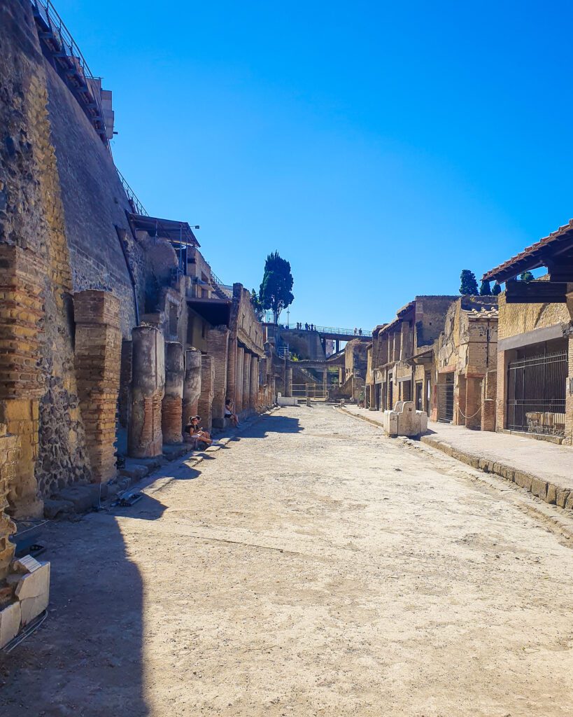 An old street in Herculaneum, Italy with stone buildings on either side and a big dirt pathway in between. The sky is blue and completely clear of any clouds above it all "See Naples On a Budget With These Six Great Ideas!"