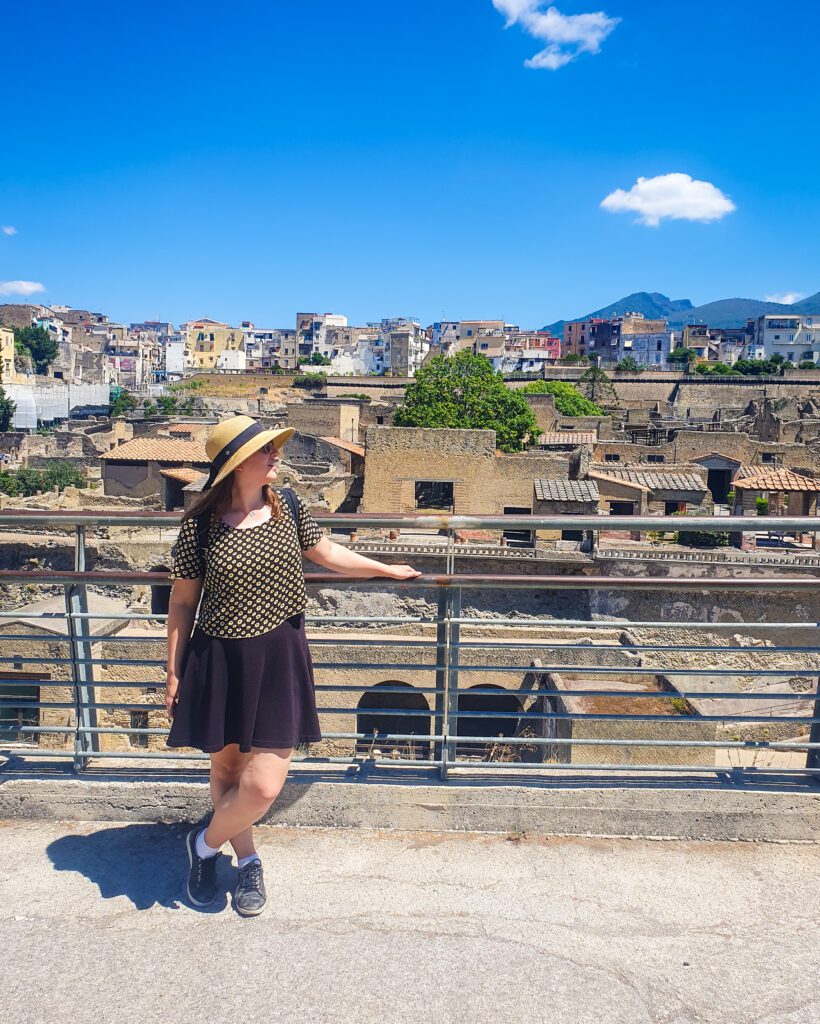 Katherine standing in front of a railing with the ruins of Herculaneum, Italy behind her and some more modern buildings and hills beyond the ruins "See Naples On a Budget With These Six Great Ideas!"