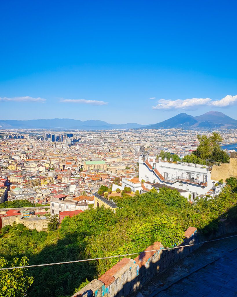 A sweeping view of Naples, Italy with its orange roofs and white buildings. Mount Vesuvius can be seen looming over the city on the right side hand side of the photo "See Naples On a Budget With These Six Great Ideas!"