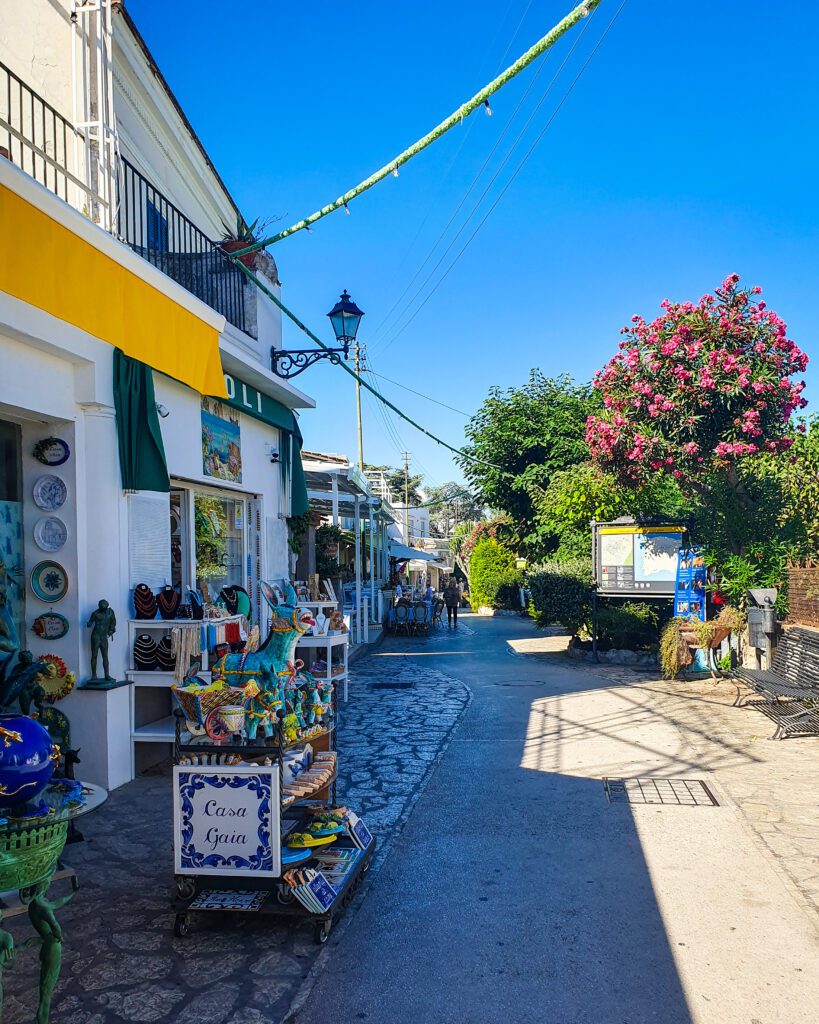 A street in Anacapri, Italy with a shop on the left with some beautiful colourful souvenirs on a tiled section of the street. Later down the street are some trees and one of them has some gorgeous pink flowers all over it "See Naples On a Budget With These Six Great Ideas!" 