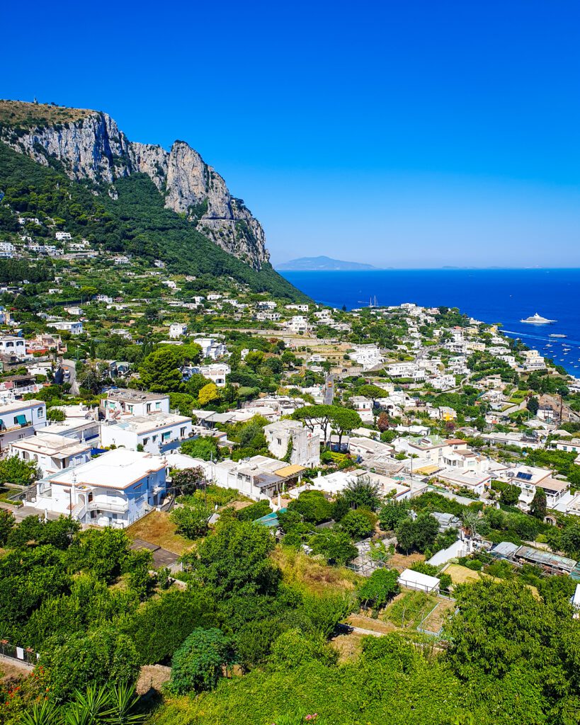 Another beautiful view over Capri, Italy with white buildings and green plants in front of a big cliff and the blue ocean "See Naples On a Budget With These Six Great Ideas!"