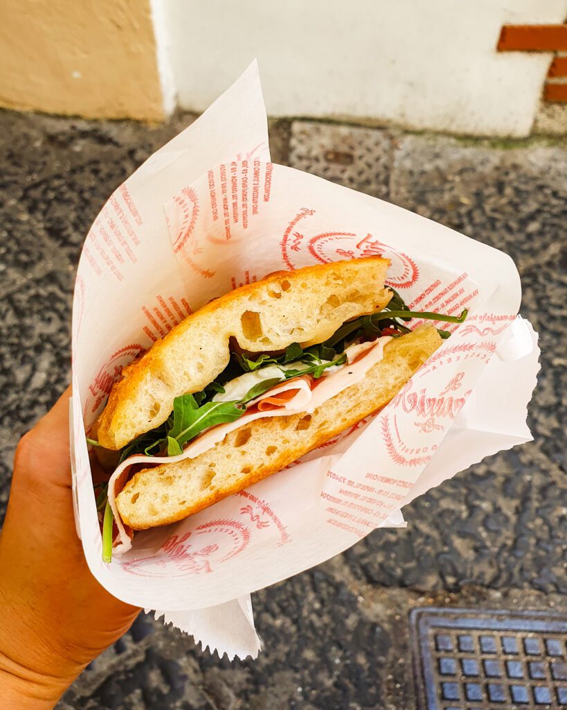 A hand holding a sandwich in paper with two pieces of focaccia and fresh greens and ham in between in Capri, Italy "See Naples On a Budget With These Six Great Ideas!"